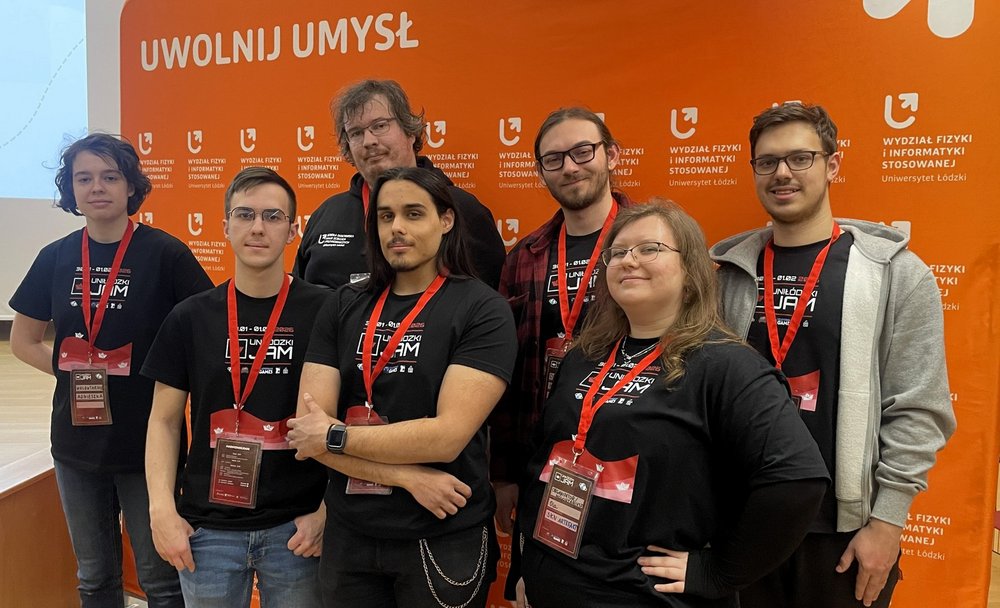 Na zdjęciu widać grupę ośmiu osób stojących obok siebie i pozujących do zdjęcia. Wszyscy mają na sobie czarne koszulki z napisem „UniŁódzki Jam 2026” oraz czerwone identyfikatory na szyjach. Stoją na tle pomarańczowej ścianki z logo Wydziału Fizyki i Informatyki Stosowanej Uniwersytetu Łódzkiego i hasłem „Uwolnij umysł”. Osoby są uśmiechnięte i wyglądają na zadowolone.