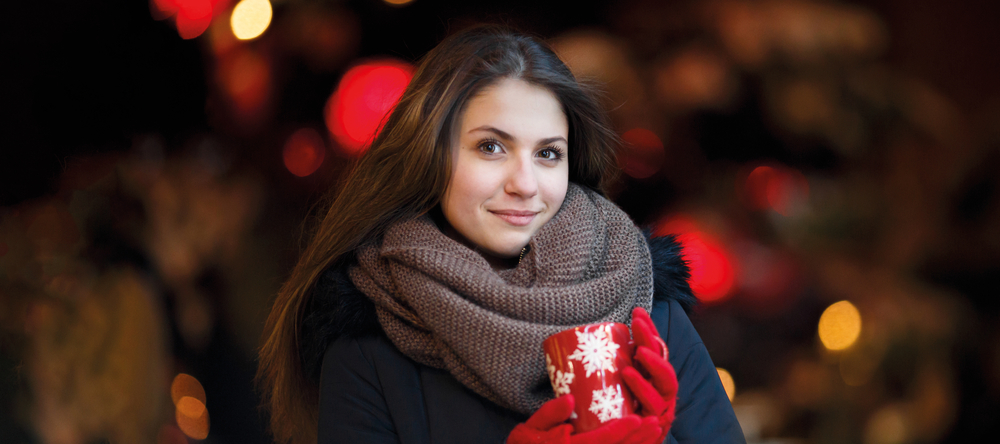 uśmiechnięta kobieta z długimi włosami ubrana w kurtkę, brązowy szalik i czerwone rękawiczki, trzymająca czerwony kubek z białymi płatkami śniegu, w tle świąteczne światła