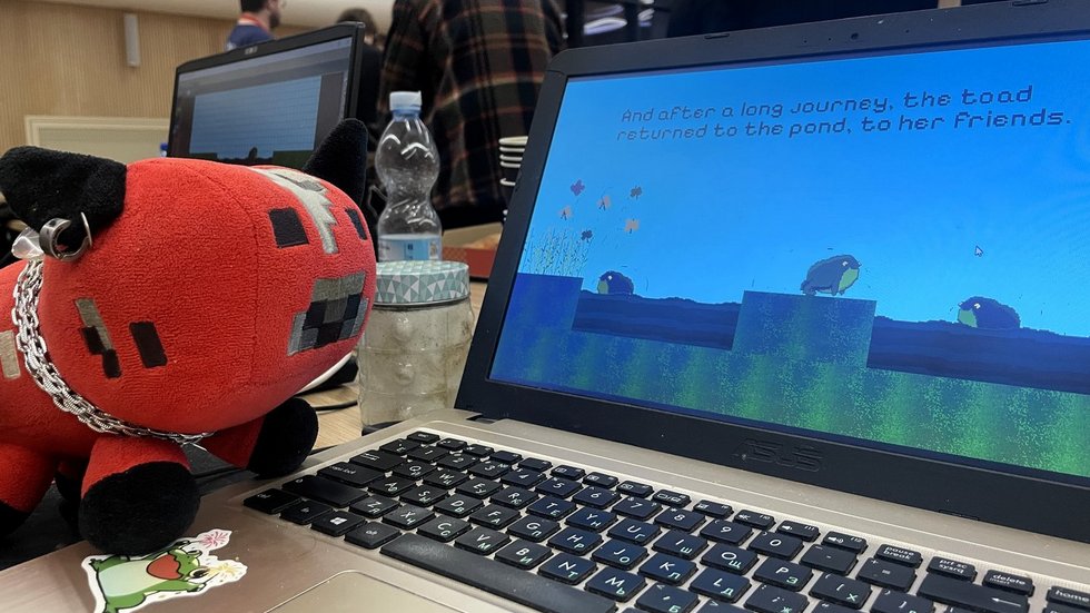 Na zdjęciu widać otwarty laptop stojący na stole podczas wydarzenia. Na ekranie wyświetlana jest gra komputerowa w stylu pikselowym z żabą na tle nieba i wody oraz tekstem końcowym gry. Obok laptopa leży czerwono-czarna pluszowa maskotka w kształcie zwierzątka z kolczykiem i łańcuszkiem. Na stole znajdują się także butelka wody i słoik. W tle widać grupę osób stojących w sali, co sugeruje, że zdjęcie zostało wykonane podczas maratonu tworzenia gier.