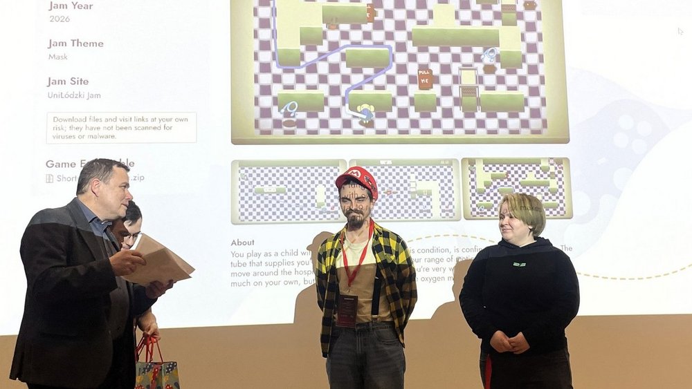 Na zdjęciu widać trzy osoby stojące przed dużym ekranem z wyświetloną prezentacją gry komputerowej zatytułowanej „Shortness of Breath”. Po lewej stronie stoi Tomasz Gwizdałła i ogłasza wyniki oraz wręcza nagrody. Pośrodku stoi młody mężczyzna w czerwonej czapce i koszuli w kratę, z identyfikatorem na szyi. Po prawej stronie stoi młoda kobieta ubrana na czarno.