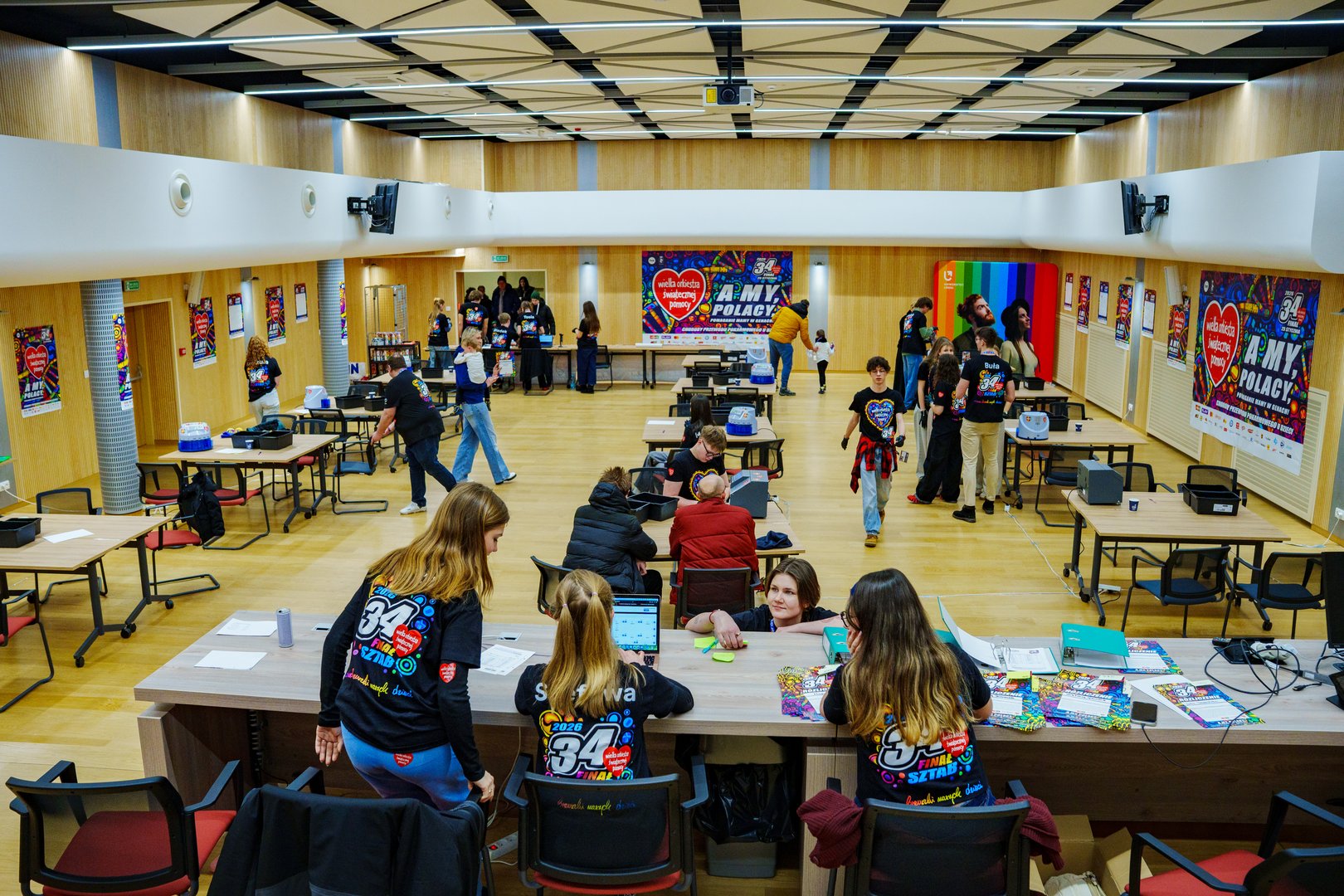 Widok pomieszczenia z ławkami i stołami i wolontariuszami- siedziby sztabu WOŚP UŁ